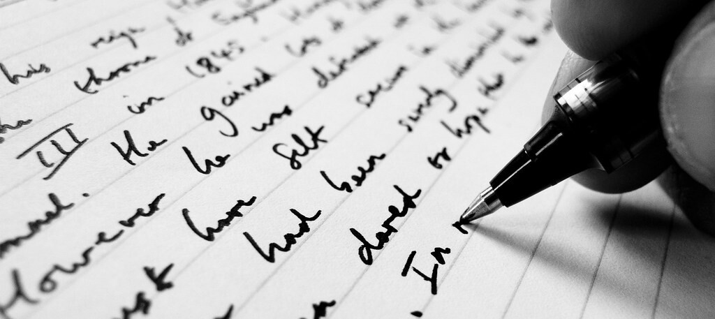 Black and white close-up of a pen writing in cursive on lined paper